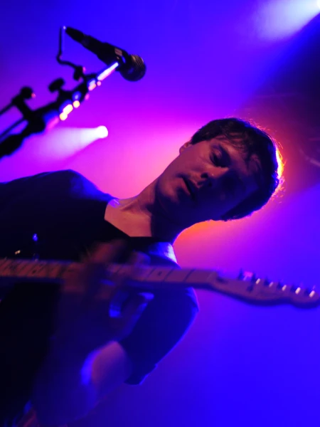 En man spelar gitarr. Rött ljus i spotlight bakom honom och blått runt om. En mic i förgrunden.