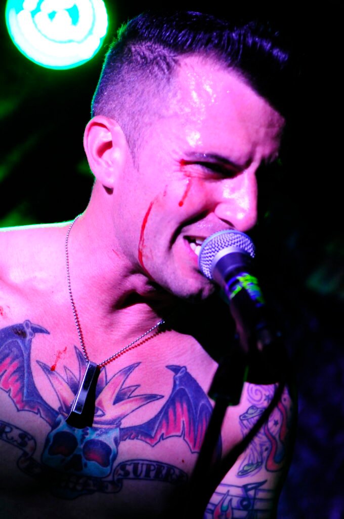 Sångaren i Koffein Kats, blöder i ansiktet framför micen, tatueringar på bar överkropp.