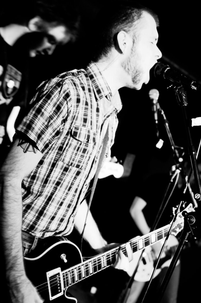 Svart-vit bild av en sångare med gitarr och rutig skjorta, han skriker i micen.
